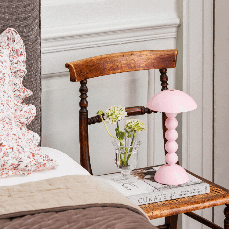 A wooden chair used as a bedside table holds the Addison Ross Bobbin LED Cordless Lamp with adjustable brightness, a small vase of white flowers, and a book, beside a bed featuring a floral pillow and beige quilt.