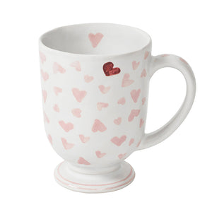 The Juliska Love You More Mug features an elegant pedestal foot, soft pink hearts, and a cherry-red heart near the rim, all on classic white ceramic.