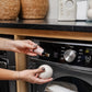 A person applies L'Avant Collective’s Laundry Oil, Blushed Bergamot, from a dropper bottle onto wool dryer balls in front of an open dryer filled with white towels.