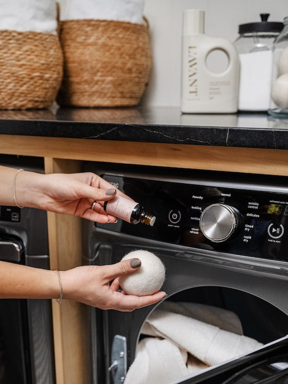 A person applies L'Avant Collective’s Laundry Oil, Blushed Bergamot, from a dropper bottle onto wool dryer balls in front of an open dryer filled with white towels.