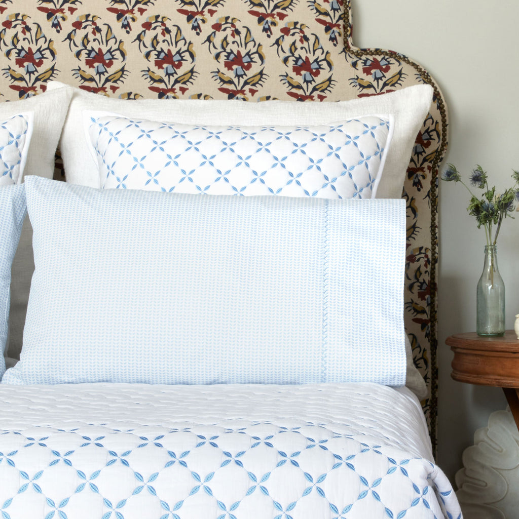 A bed features the John Robshaw Cinde Bedding Collection in Light Indigo, with a decorative headboard, a small wooden side table holding a vase of flowers, and white pillows decorated with blue hand embroidery.