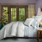 A neatly made bed is dressed in the John Robshaw Layla Bedding Collection, featuring patterned white and blue bedding with a soft cotton voile quilt inspired by Mughal gardens. The bed sits beside a wooden nightstand and large glass doors overlooking a green outdoor area.