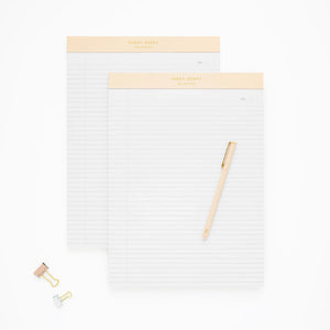 Two Sugar Paper Legal Pads with beige headers, reminiscent of a classic Legal Pad, are arranged on a white background alongside a beige pen resting on one pad and two binder clips—an ideal setup for organizing your to-do list or bringing sophisticated style to any professional desk.