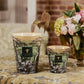 Two Baobab Collection Sacred Trees Touba Candles by Baobab are lit and placed on a patterned surface beside a vase of assorted flowers, with a brick wall and window in the background.
