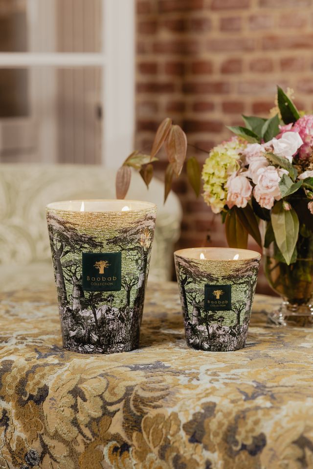 Two Baobab Collection Sacred Trees Touba Candles by Baobab are lit and placed on a patterned surface beside a vase of assorted flowers, with a brick wall and window in the background.