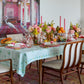A dining table set with a Kim Seybert Lima Seafoam Tablecloth, floral centerpieces, pink candles, fruits, glassware, and wooden chairs, with a colorful ornate room photo on the wall behind.