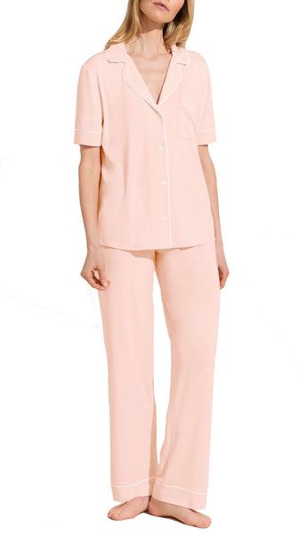 Woman standing in a light pink, short-sleeved Eberjey Gisele Short Sleeve Long Pant PJ Set with white piping, made from sustainable TENCEL™ Modal fabric, consisting of a button-up top and matching pants.