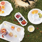 A picnic scene on grass features trays of sliced citrus, berries, drinks in glasses, and a straw hat with a black ribbon, all artfully served on Juliska’s Berry & Thread Melamine Whitewash Dinnerware and Serveware Collection for easy outdoor dining.