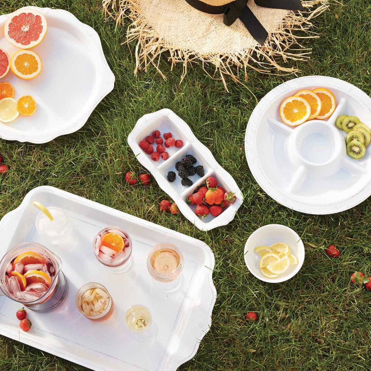 A picnic scene on grass features trays of sliced citrus, berries, drinks in glasses, and a straw hat with a black ribbon, all artfully served on Juliska’s Berry & Thread Melamine Whitewash Dinnerware and Serveware Collection for easy outdoor dining.