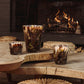 Three hand-blown Baobab Collection Halona Yellow World candles with an abstract brown and yellow pattern sit on a live-edge wooden coffee table in front of a lit fireplace.