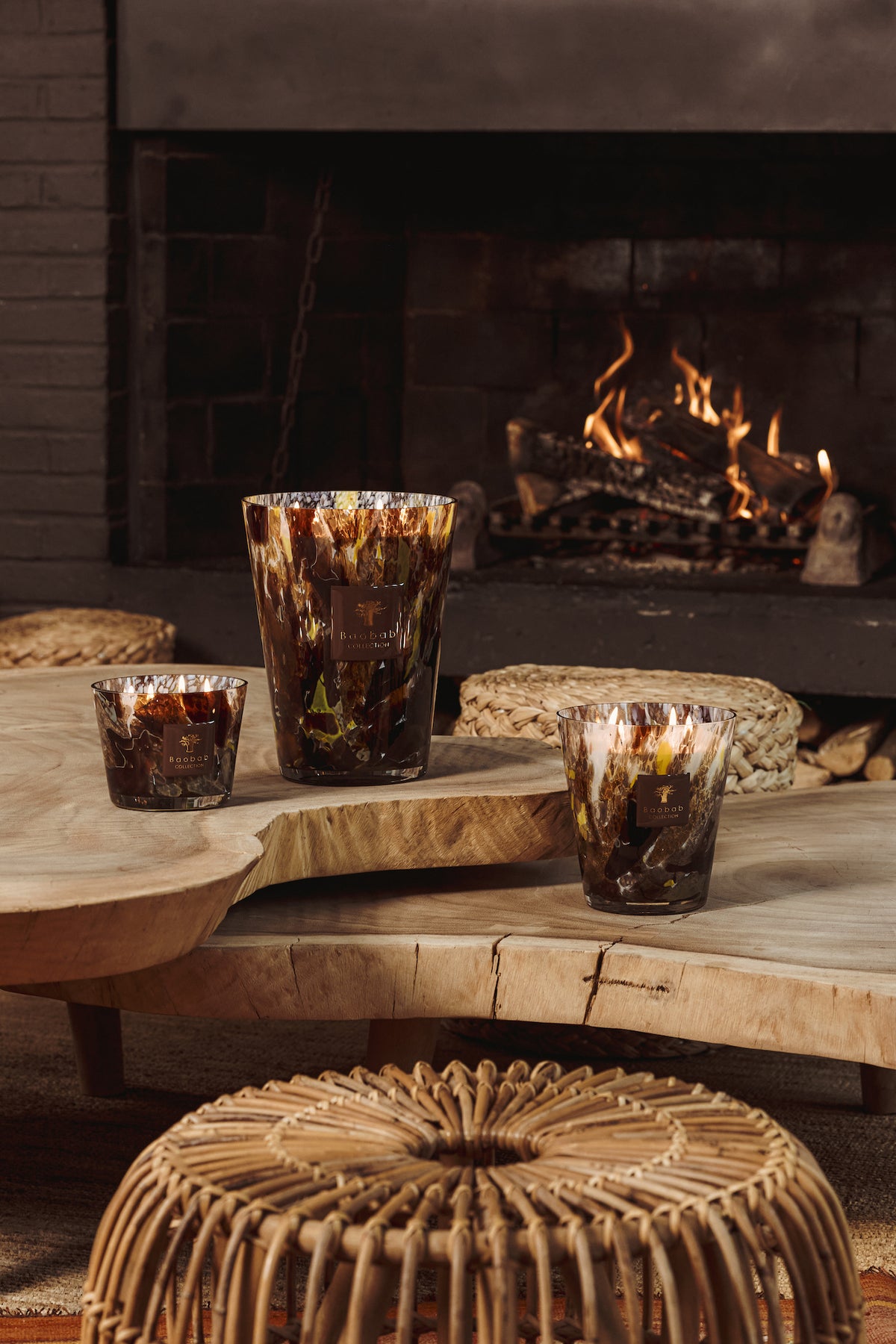 Three hand-blown Baobab Collection Halona Yellow World candles with an abstract brown and yellow pattern sit on a live-edge wooden coffee table in front of a lit fireplace.