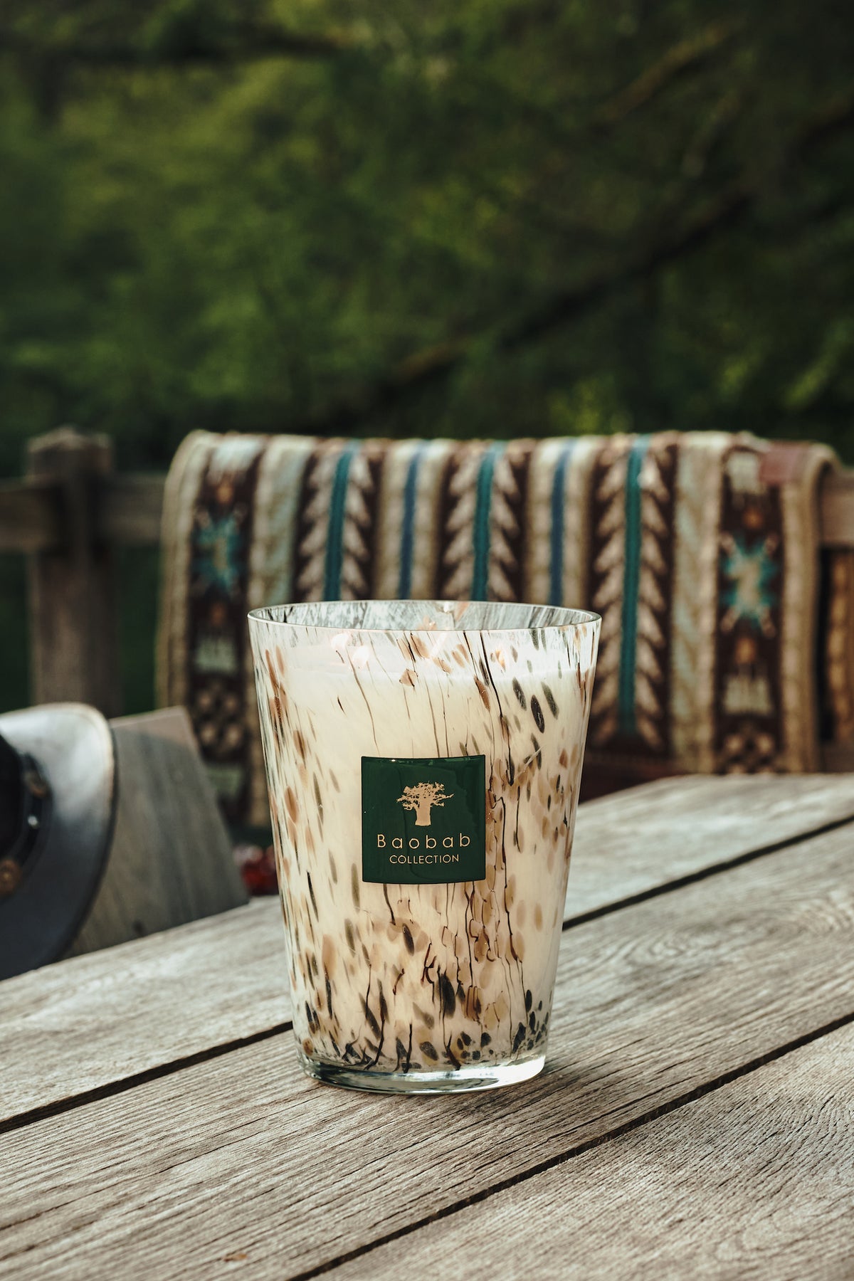 Baobab Collection Halona White World Candle, a hand-blown glass candle holder with a brown and white marbled pattern and a square logo label, sitting on a wooden table.