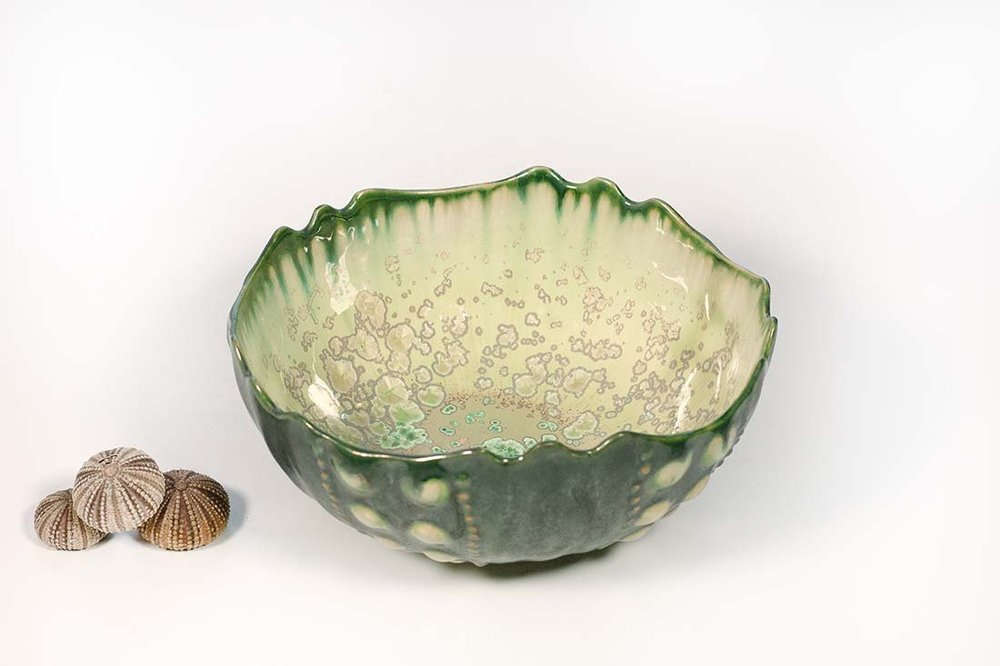 A green wavy-rimmed bowl from AE Ceramics’ handmade Sea Urchin Bowl Collection sits beside three small seashells on a white background.