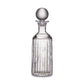 The Montpelier Drinkware Collection by Simon Pearce features a clear hand-blown glass decanter with vertical etched lines, a round stopper, and a narrow neck, set against a plain white background.