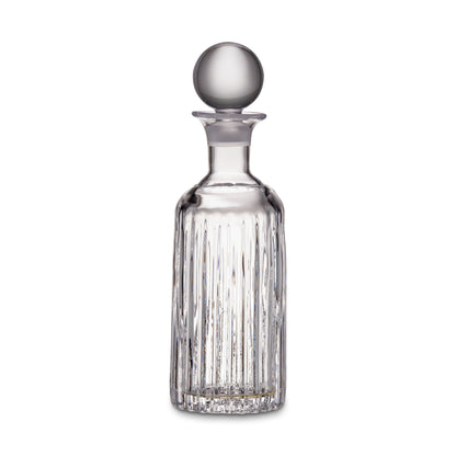 The Montpelier Drinkware Collection by Simon Pearce features a clear hand-blown glass decanter with vertical etched lines, a round stopper, and a narrow neck, set against a plain white background.