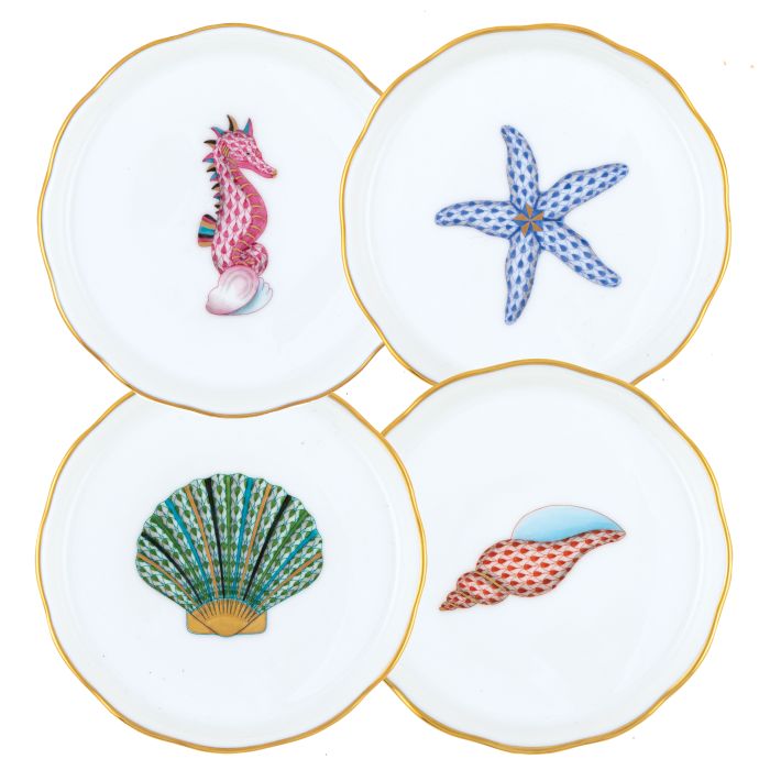 The Herend Aquatic Coaster Set includes four durable porcelain coasters, each with a gold rim and a unique hand-painted sea-themed design: pink seahorse, blue starfish, green seashell, and red conch shell.