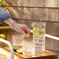 A person places a lime wedge into a Simon Pearce Montpelier Drinkware Collection glass beside a pitcher with lime slices, two glasses, and cut limes on a wooden tray outdoors.