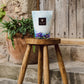 A blue and white vase with flowers sits on a wooden stool, accompanied by the Baobab Collection Eden Seaside Candle by Baobab nearby. In the background, a green plant in a terracotta pot adds charm against the rustic wooden door.