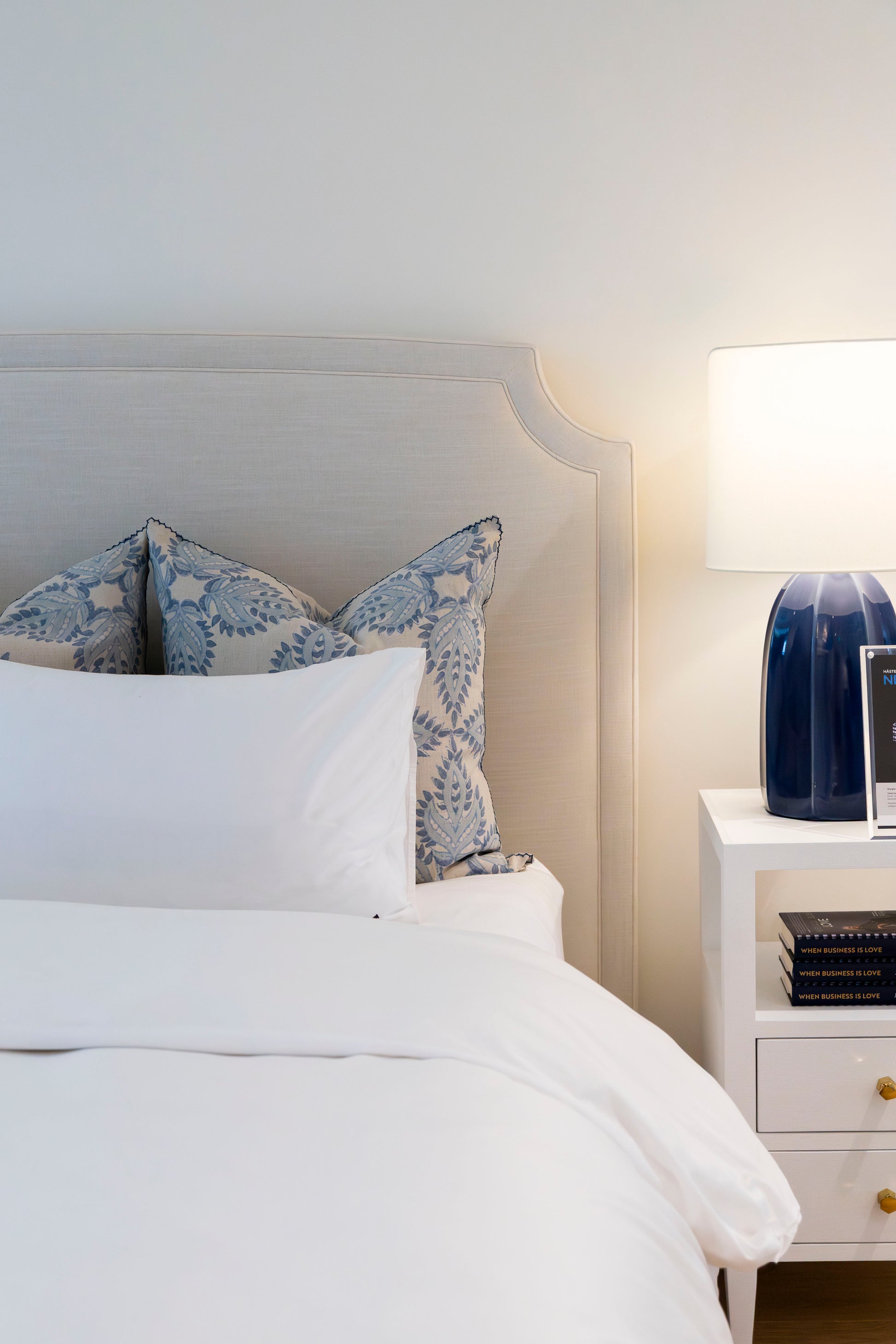 A neatly made bed with white linens and blue-patterned pillows showcases the Lee Industries Laguna Cloud Cut Corner King Bed, paired with a white nightstand, lamp, and stacked books for a touch of contemporary elegance.