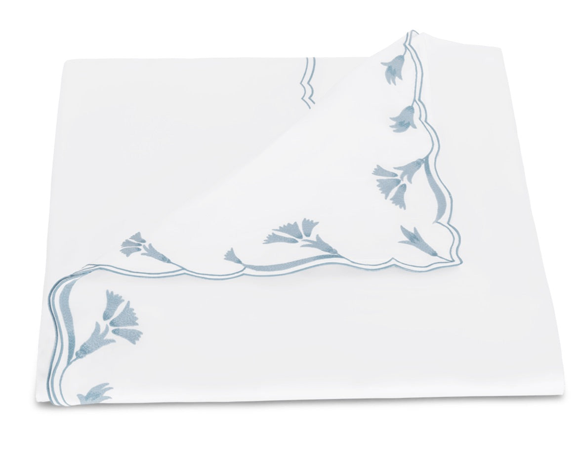 A folded white napkin from the Matouk Marcella Bedding Collection, featuring a blue scalloped floral border along two edges reminiscent of embroidered bedding, displayed on a white background.