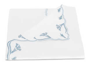 The Matouk Marcella Bedding Collection in Hazy Blue features a white cloth napkin with scalloped edges and delicate blue floral embroidery along the border, displayed folded at one corner.