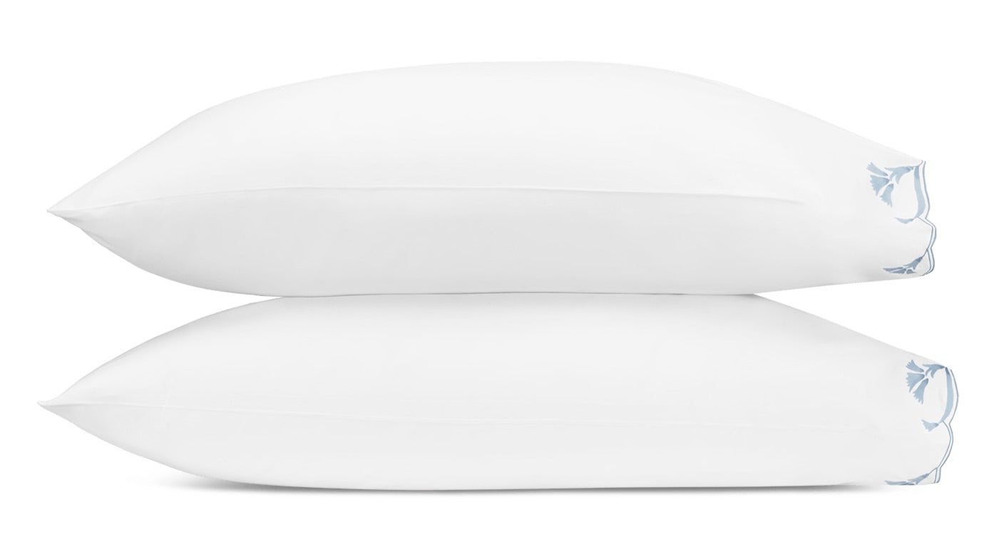 Two white pillows from the Matouk Marcella Bedding Collection are stacked, each pillowcase featuring a blue scalloped floral pattern on the bottom right corner.