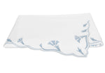 A folded white cloth napkin with scalloped edges and delicate light blue floral embroidery in one corner, echoing the signature scalloped florals of Matouk’s Marcella Bedding Collection.