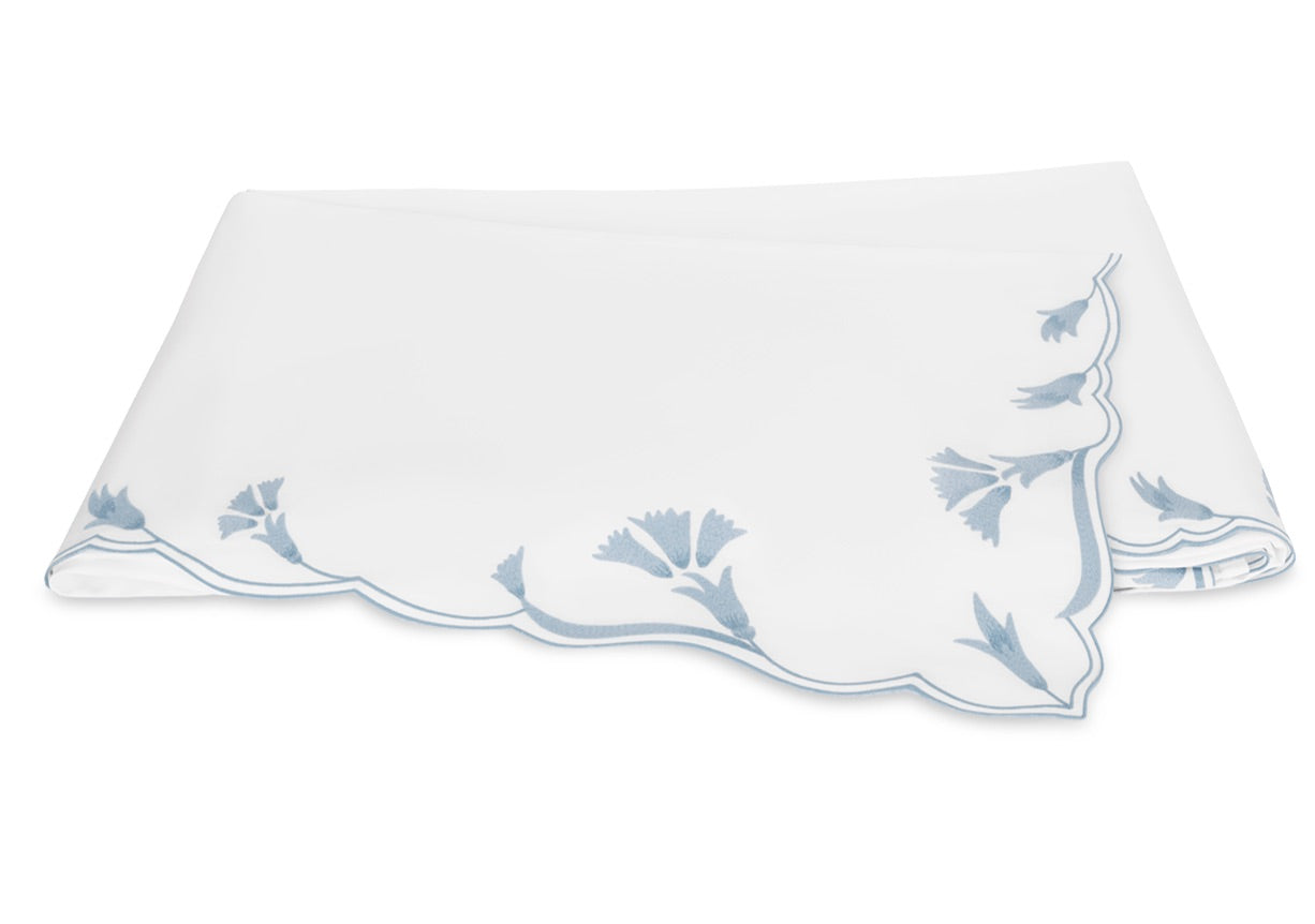 A folded white cloth napkin with scalloped edges and delicate light blue floral embroidery in one corner, echoing the signature scalloped florals of Matouk’s Marcella Bedding Collection.