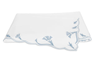 A folded white cloth napkin with scalloped edges and delicate light blue floral embroidery in one corner, echoing the signature scalloped florals of Matouk’s Marcella Bedding Collection.