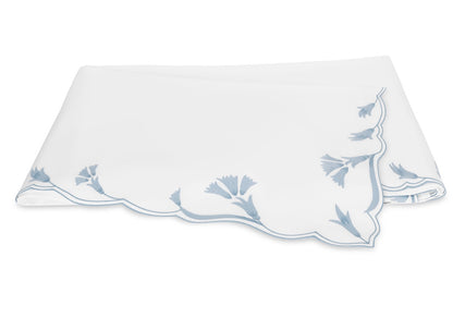 A folded white cloth napkin with scalloped edges and delicate light blue floral embroidery in one corner, echoing the signature scalloped florals of Matouk’s Marcella Bedding Collection.