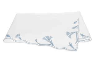 The Marcella Bedding Collection by Matouk in Hazy Blue features elegant scalloped edges and blue floral embroidery on a crisp white background.