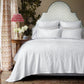 A neatly made bed is dressed with the Meera Bedding Collection from John Robshaw—featuring white bedding and a woven cotton coverlet that highlights artisanal craftsmanship inspired by Portuguese textile traditions. The space also includes a patterned headboard, a round side table with flowers and a lamp, and curtains with a geometric design.