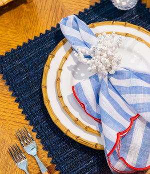 A white plate rests on a navy placemat. The Amalfi Napkin Set by Kim Seybert, with blue and white stripes and red trim, is held by a coral-shaped napkin ring. Two forks beside the setting evoke a Mediterranean lifestyle.
