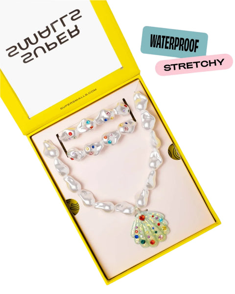 The Supersmalls Mermaid Pool Party Mega Set includes mermaid-themed jewelry for kids, featuring a pearl necklace with a vibrant, gem-encrusted seashell pendant and a matching bracelet. The set is beautifully displayed in an open yellow box labeled "Super Smalls" and comes with tags that read "Waterproof" and "Stretchy," making it the perfect accessory for the beach.