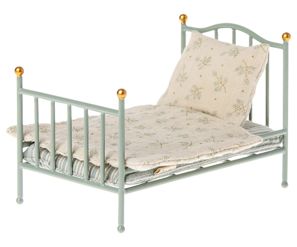 A small, mint-colored vintage metal bed frame from Maileg, complete with a pillow and a folded bedspread featuring a subtle floral pattern, perfect for your Maileg friends.