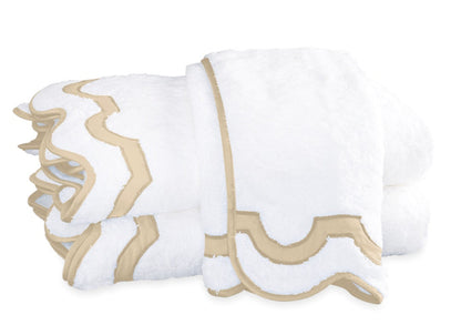 Two folded white towels with beige scalloped trim from the Matouk Mirasol Bath Collection are stacked on a white surface.