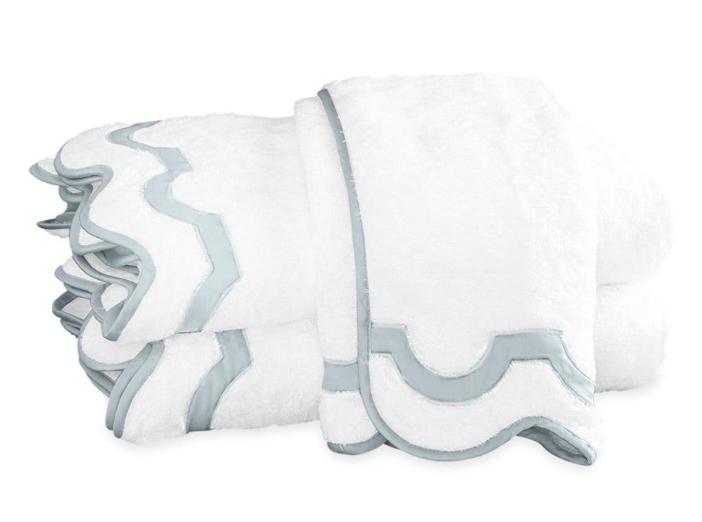 A stack of folded Mirasol Bath Collection towels by Matouk, crafted from white Cairo cotton terry with light blue trim, arranged on a plain white background.