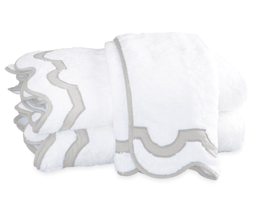 Two folded white towels from the Matouk Mirasol Bath Collection feature decorative scalloped edges trimmed in light gray and are stacked on a white background.