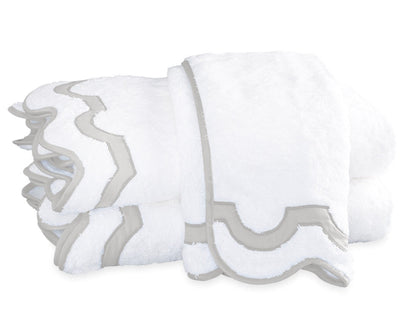 Two folded white towels from the Matouk Mirasol Bath Collection feature decorative scalloped edges trimmed in light gray and are stacked on a white background.