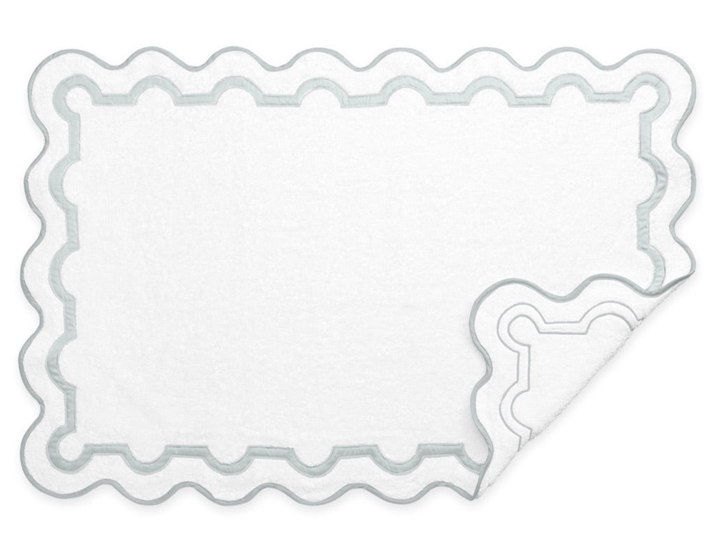 White rectangular bath mat with scalloped light grey borders and a folded corner, perfectly complementing Matouk’s Mirasol Bath Collection.