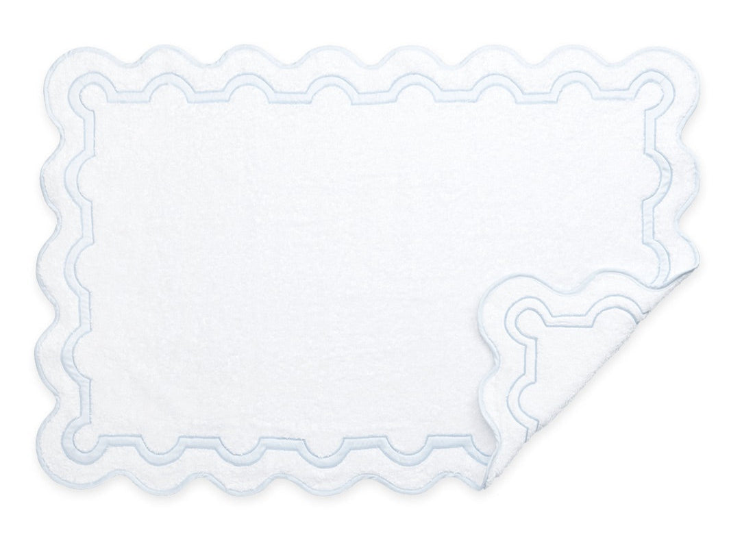 White rectangular bath mat from the Matouk Mirasol Bath Collection featuring scalloped edges and a blue wavy border. One corner is folded to show the reverse side.