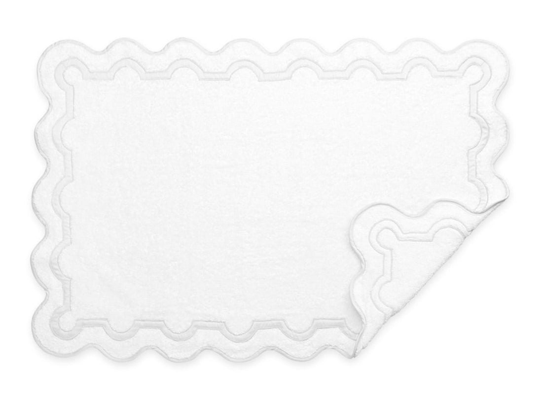 A white rectangular bath mat with scalloped edges and a raised border, inspired by the Matouk Mirasol Bath Collection. One corner is slightly folded over, showcasing the elegant Matouk design details.