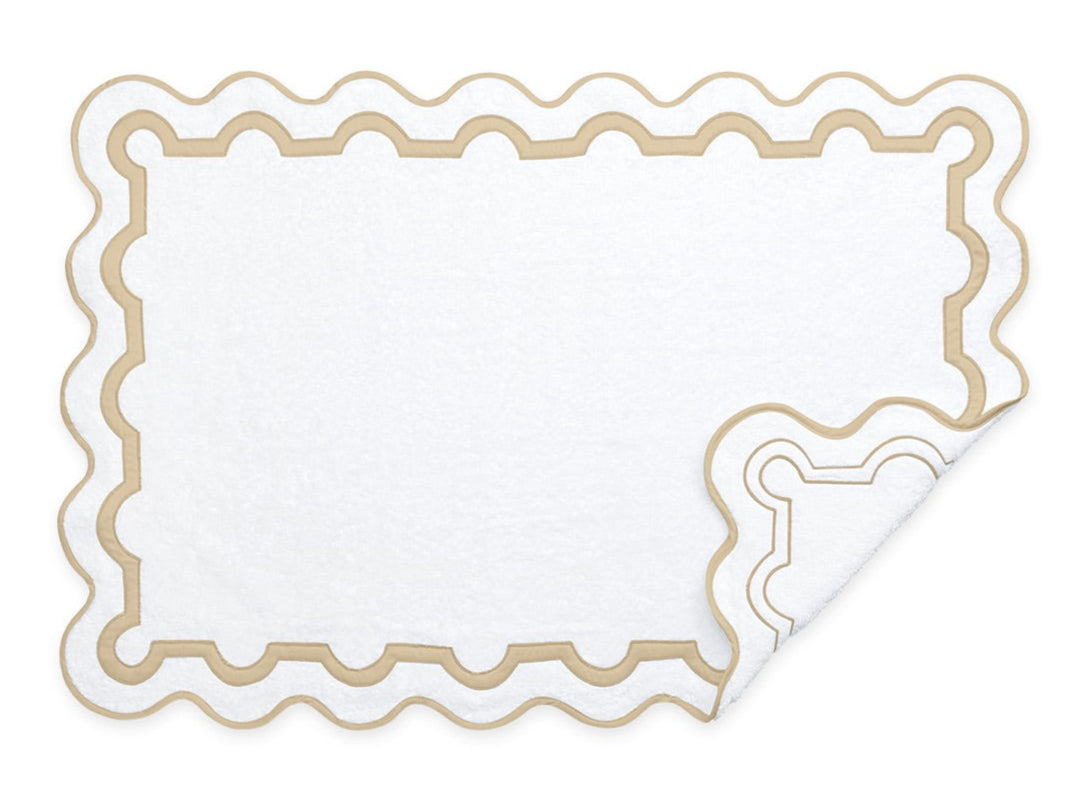 The Matouk Mirasol Bath Collection features a white rectangular bath mat with a scalloped beige border, folded corner, and elegant reversible pattern, designed to coordinate with Matouk’s Cairo cotton terry towels.