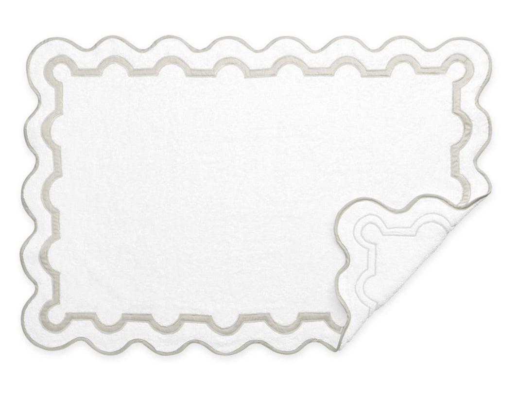 Matouk’s Mirasol Bath Collection features a white rectangular bath mat with scalloped edges and a contrasting border, shown with one corner flipped—pairs perfectly with Cairo cotton terry towels and other Matouk Mirasol pieces.
