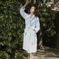A woman in a white Matouk Milagro Robe, made from OEKO-TEX certified cotton, stands barefoot outdoors holding flowers, surrounded by lush greenery and trees.
