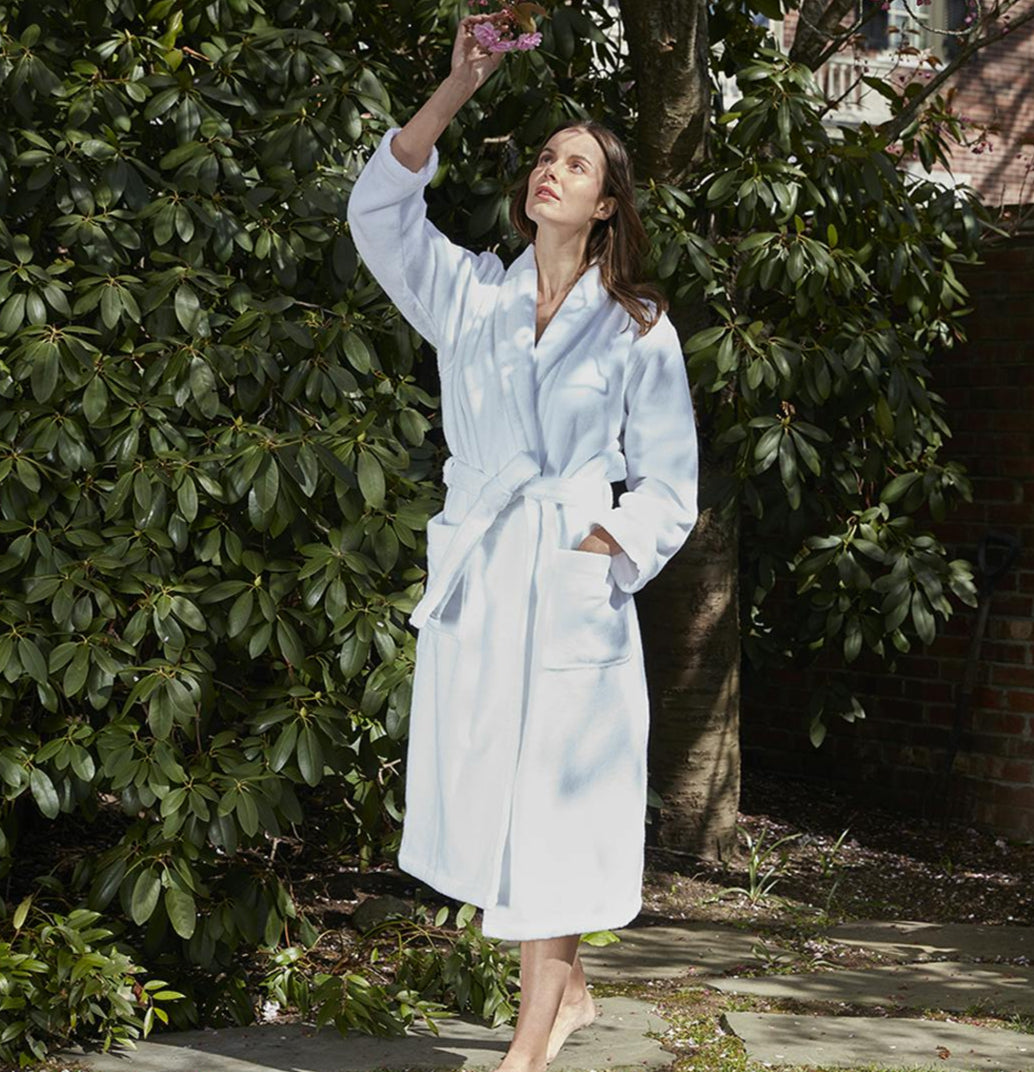 A woman in a white Matouk Milagro Robe, made from OEKO-TEX certified cotton, stands barefoot outdoors holding flowers, surrounded by lush greenery and trees.