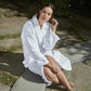 A woman in a white Matouk Milagro Robe sits on a stone surface near water, one hand on her knee and the other touching her hair.