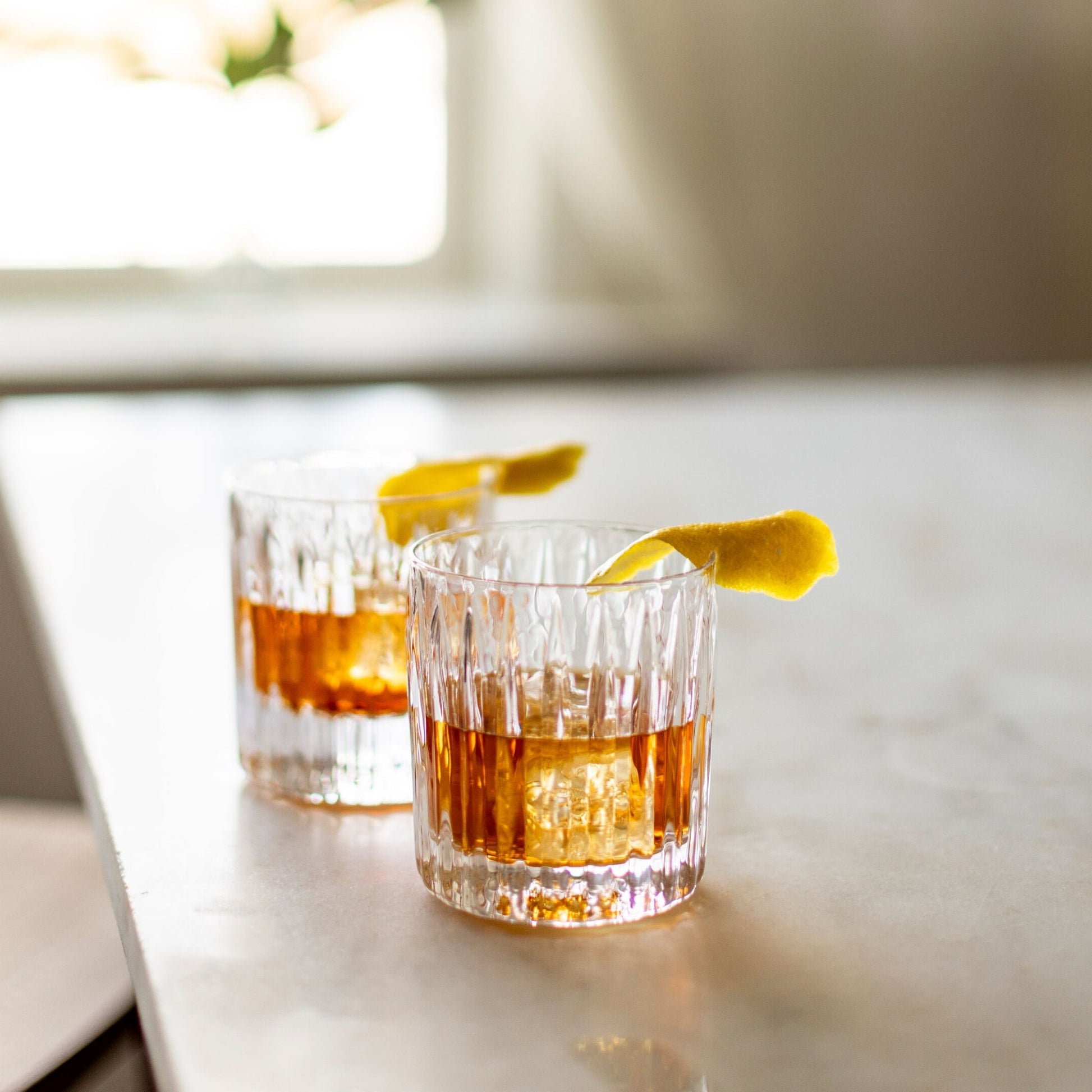 Two hand-blown double old-fashioned glasses from the Montpelier Drinkware Collection by Simon Pearce hold amber cocktails with large ice cubes and lemon twists, set on a light-colored surface.