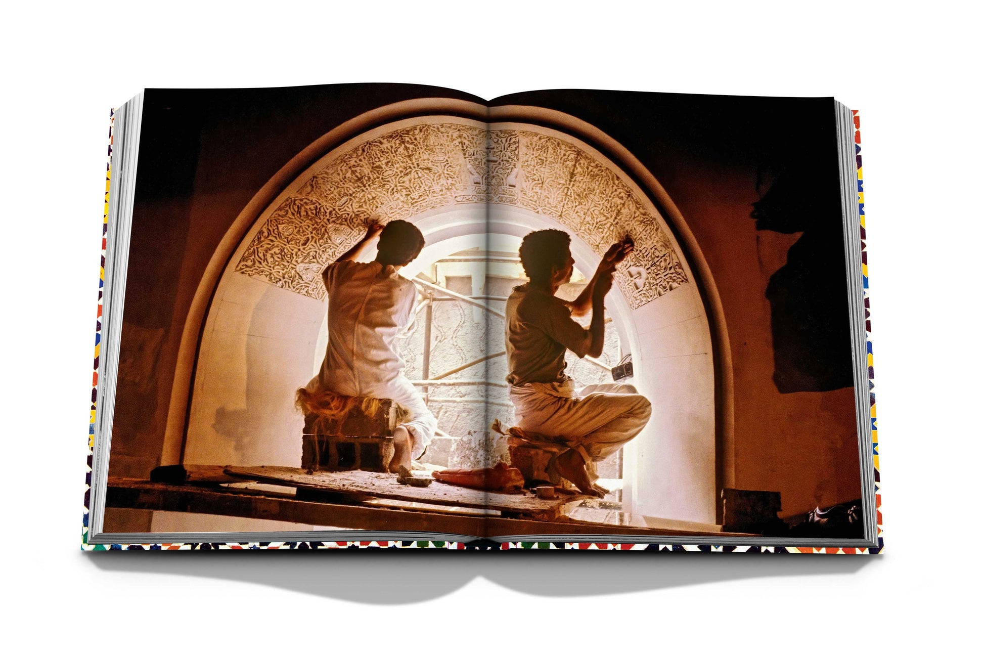 Two individuals, showcasing exceptional craftsmanship, are working on an intricate archway inspired by Assouline's Moroccan Decorative Arts, as seen in an open book with a decorative peripheral border designed by Philippe Saharoff.
