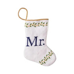 The Bauble Stockings Mr. Needle Point Stocking features "Mr." embroidered in blue on white fabric, with green leaf designs and a gold loop—ideal for a wedding gift or festive mantle decor.